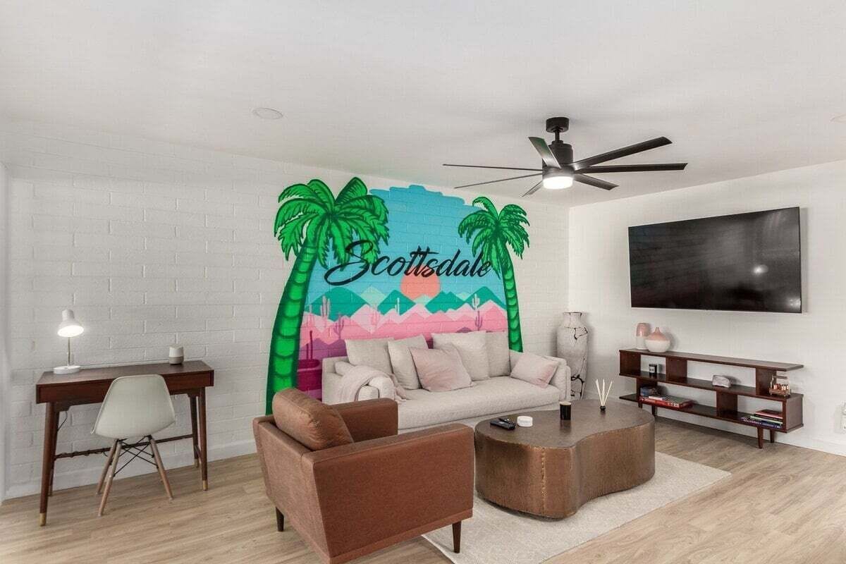 A modern living room with a white brick wall, a large flat-screen TV, a brown sofa, a coffee table, and a painting of palm trees and mountains.