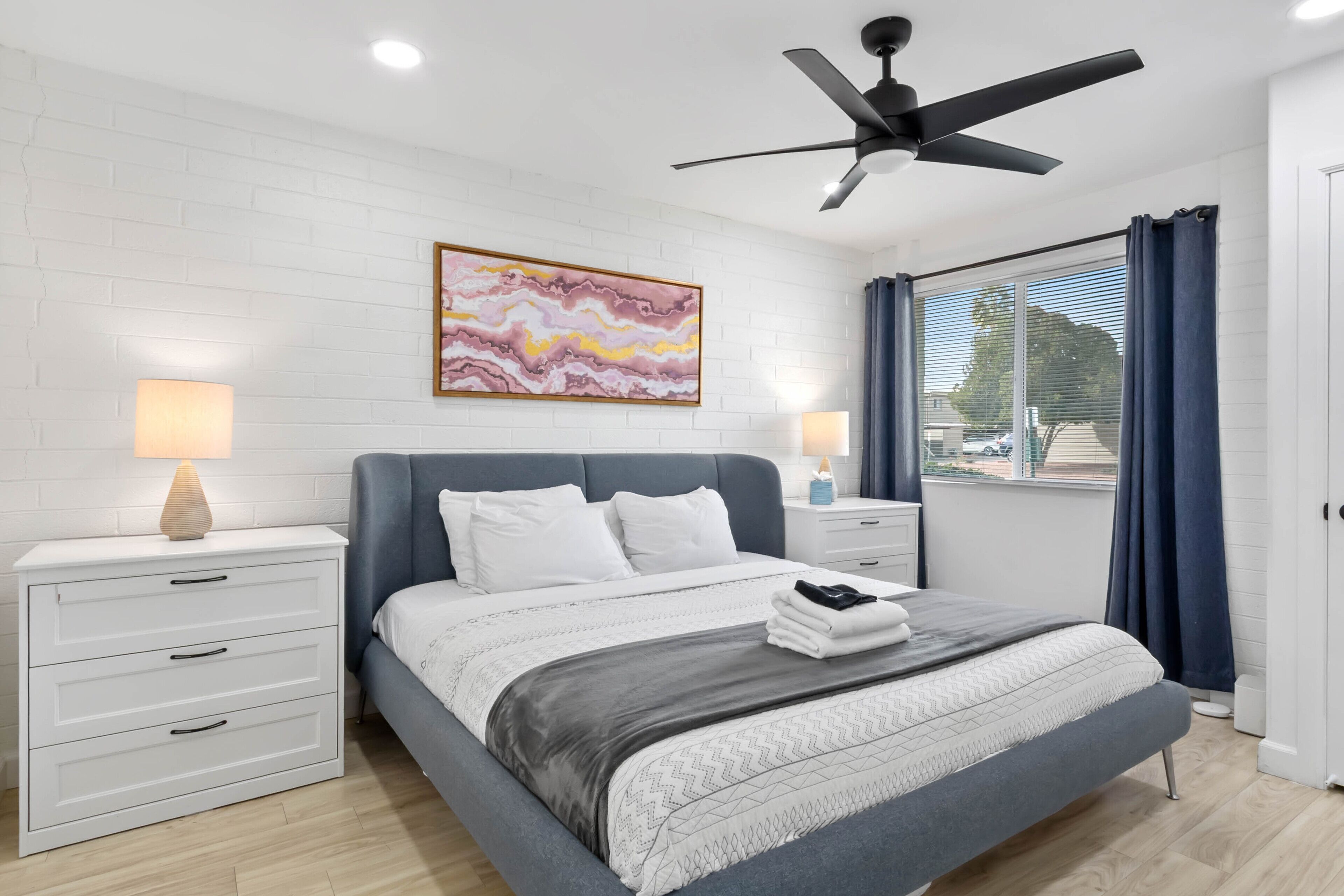 A modern bedroom with a bed, nightstands, a ceiling fan, and a large window with curtains.