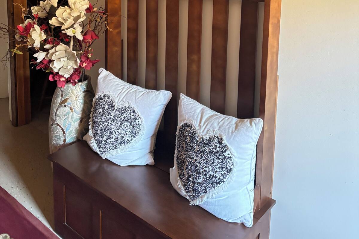 A wooden bench with heart-patterned pillows, a welcome sign, and a wicker basket on a wall.