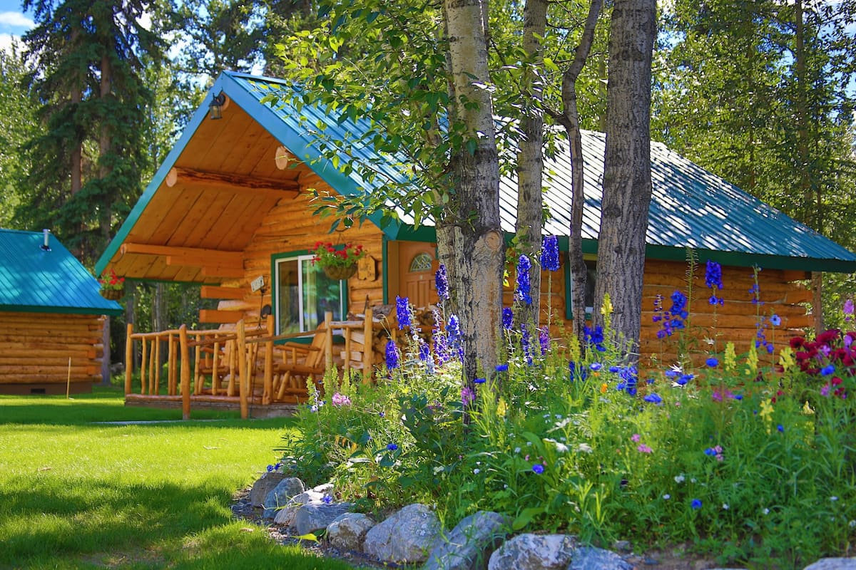 A log cabin with a green roof, wooden deck, and surrounding garden with various flowers.