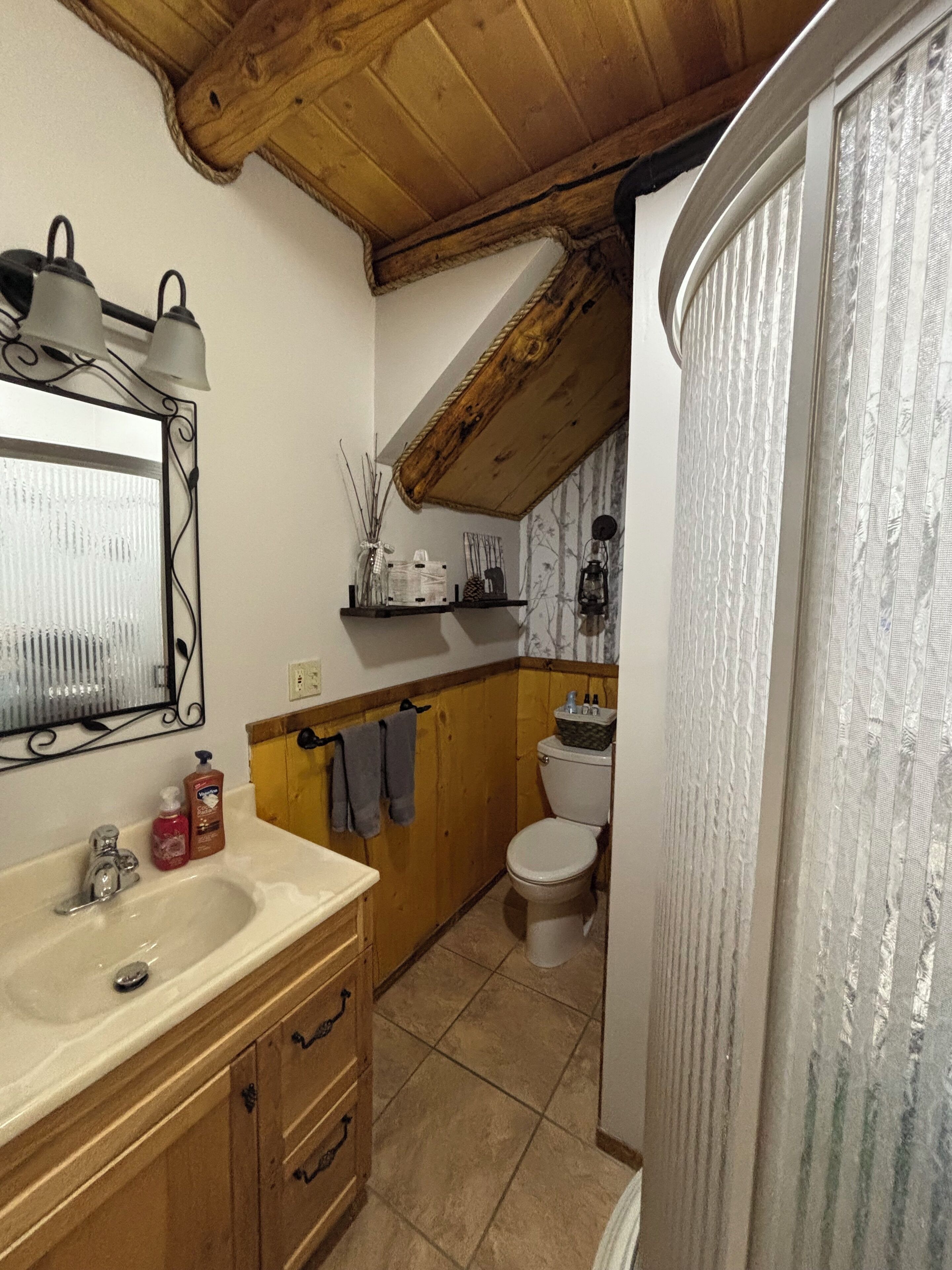A bathroom with a wooden ceiling, a sink with toiletries, a toilet, and a mirror with a decorative frame.