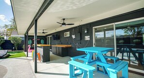Een terras buiten met een blauwe picknicktafel, een barbecue en een plafondventilator.