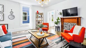 A modern living room with a blue sofa, orange armchairs, a wooden coffee table, and a TV mounted on a wooden cabinet. - Walk to BeltLine, Stylish 2BR w/ Deck, Sleeps 4 (Atlanta)