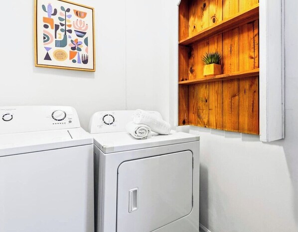 A laundry room with two white washing machines, a wooden shelf with a plant, and a framed abstract art piece on the wall.