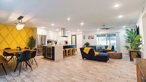 Eine moderne Küche mit weißen Schränken, einer Kochinsel und Barhocker. Ein Essbereich mit Holztisch und Stühlen lädt zum Verweilen ein. Das Wohnzimmer zeichnet sich durch ein dunkles Sofa und einen Couchtisch aus.