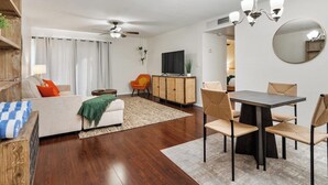 Una sala de estar moderna con un sofá, una mesa de comedor y sillas. Se ve un televisor y un aparador al fondo.