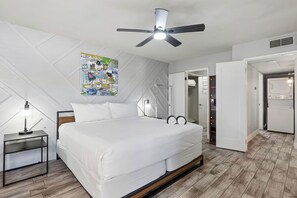 Un dormitorio con cama, ventilador de techo, mesita de noche, un cartel en la pared y una puerta que da a otra habitación.