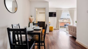 A modern living room with a dining area, a flat-screen TV mounted on the wall, and a view of the outdoors through large windows. - Canary Wharf Gem - Prvt Parking - Fast WiFi - O2 & Excel - Sleeps 4 (London)