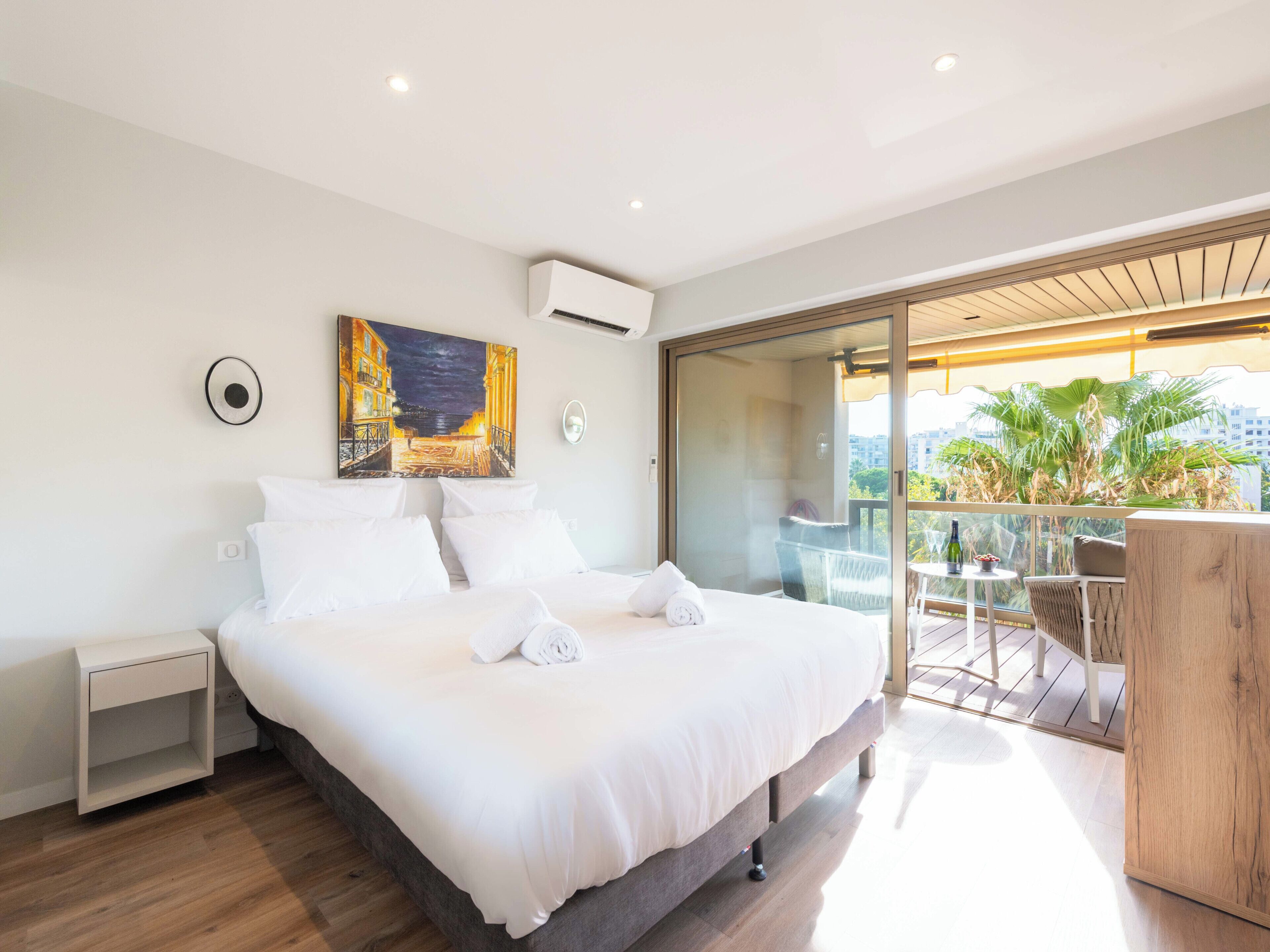 Une chambre d’hôtel moderne avec un grand lit, un tableau, un appareil de climatisation, une table de chevet et un balcon offrant une vue sur la ville.