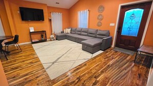 Una sala de estar con un sofá seccional, una mesa de comedor con sillas, un televisor de pantalla plana y piso de madera.