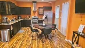 Una cocina con armarios oscuros, un refrigerador de acero inoxidable, una estufa y una cafetera. Hay una mesa de comedor con sillas y un cubo de basura.