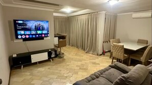 A living room with a flat-screen TV, a sofa, a dining table, and a curtain-covered window.