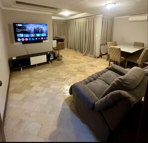 Una sala de estar con una televisión de pantalla plana, un sofá, una mesa de comedor y una ventana cubierta con cortinas.