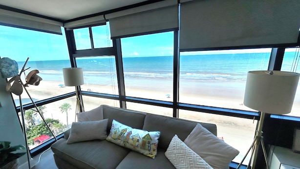A modern room with a large window overlooking a beach, a sofa with patterned cushions, and a lamp on a side table. - Spectacular views - direct oceanfront studio w/kitchen & pool (Daytona Beach)