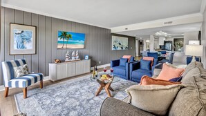 Un salon moderne avec une table basse en verre, un fauteuil à rayures bleues et grises et un canapé gris agrémenté de coussins orange.