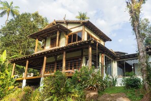 A two-story wooden house with large windows and a balcony, surrounded by lush greenery.