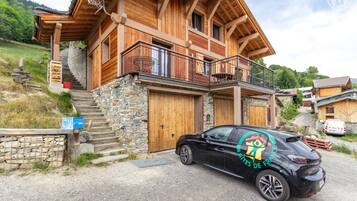 Une maison en bois à deux étages, avec des fondations en pierre, un balcon et une voiture garée devant.