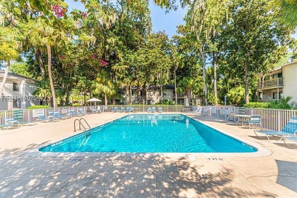 A rectangular swimming pool surrounded by lounge chairs and a fence, with trees and buildings in the background.
