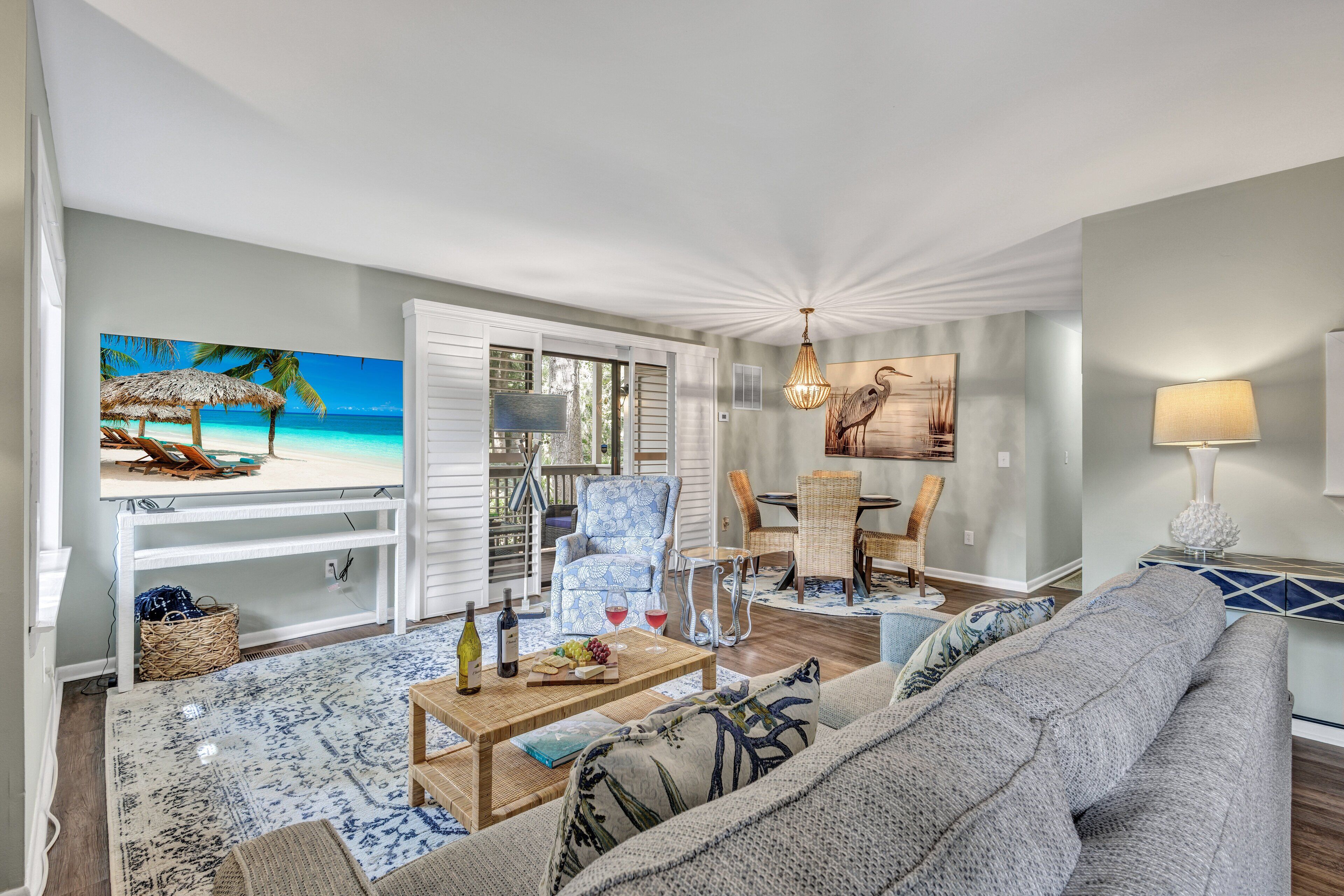 A living room with a beach-themed painting, a wicker chair, a patterned sofa, and a dining area with a table and chairs.
