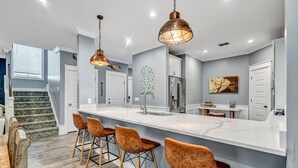 Una cocina moderna con una isla de mármol, taburetes de bar, luces colgantes y un refrigerador de acero inoxidable.
