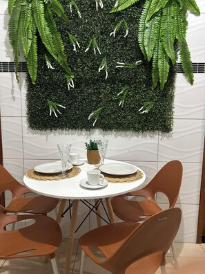 A dining area with a round table set for two, surrounded by brown chairs. A decorative wall with green foliage and white flowers is behind the table.