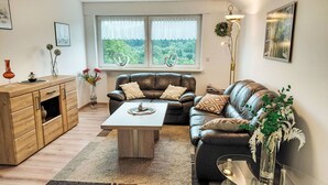 Ein modernes Wohnzimmer mit einer Ledersofa, einem Holzschrank, einem Glastisch und einem großen Fenster mit Blick auf Grünflächen.