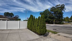 A suburban street with a white fence, a row of evergreen trees, and a clear blue sky. - Sunny House in Queens · Entire Home · Private Parking (Queens)