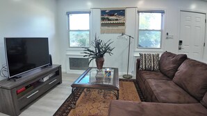 A living room with a flat-screen TV on a wooden stand, a glass coffee table with a plant, a brown leather sofa, and a patterned rug. - Sunny House in Queens · Entire Home · Private Parking (Queens)