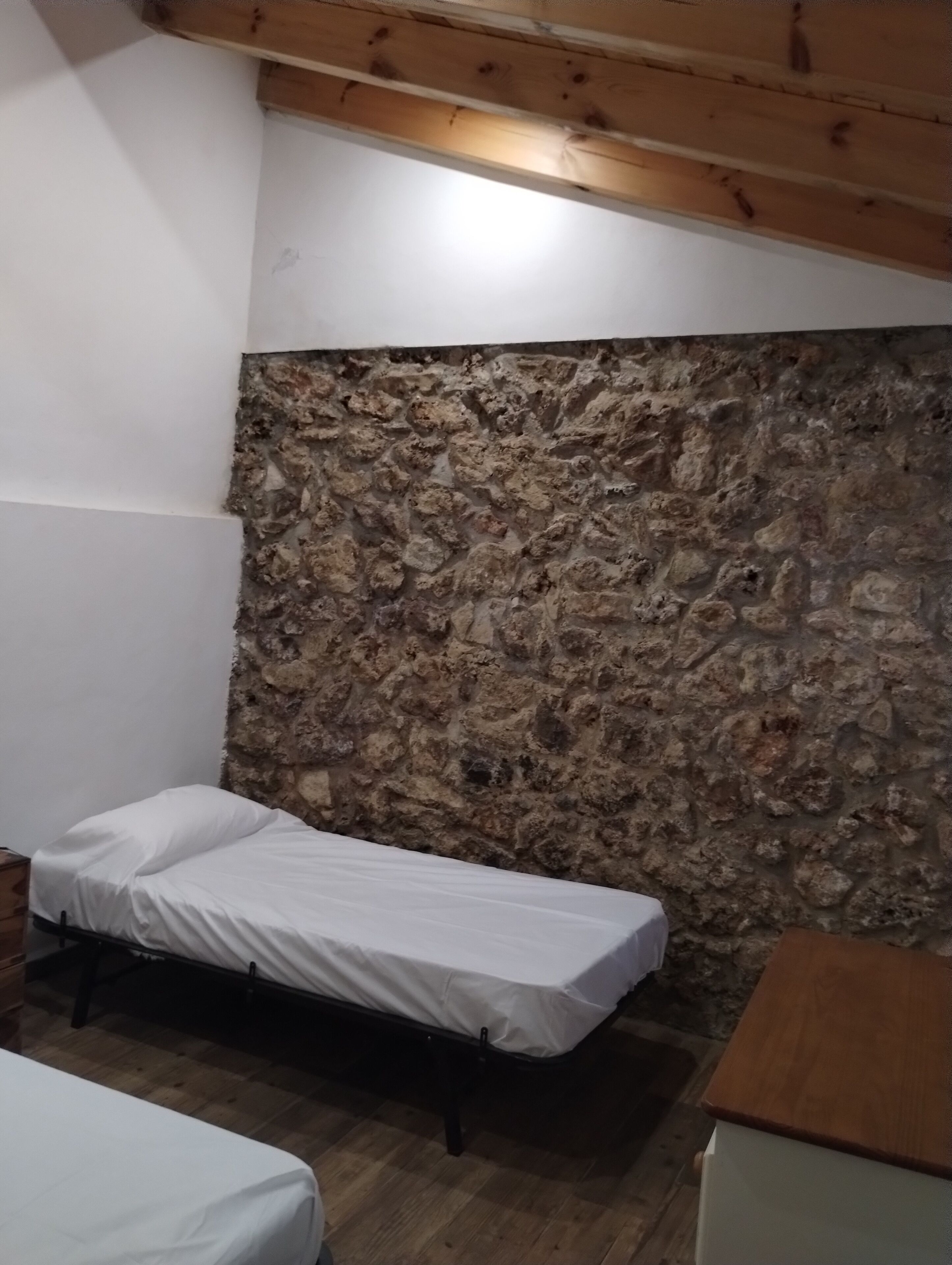 A small, single-bed room with a stone wall and wooden ceiling beams.
