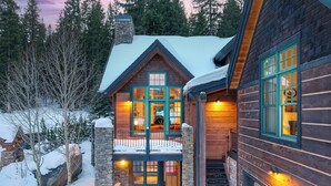 A log cabin with a stone chimney, a hot tub, and a porch with a stone railing.