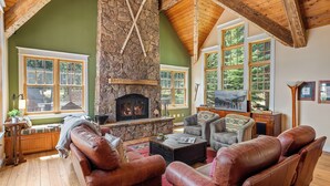 A cozy living room with a stone fireplace, leather sofas, and large windows offering a view of the outdoors.