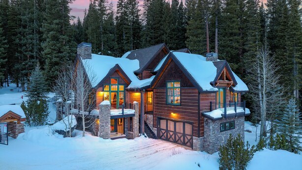 A log cabin with a stone chimney, multiple windows, and a snow-covered landscape.