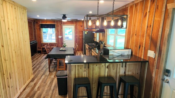 A wooden interior with a kitchen area featuring a bar counter and stools, a dining table with chairs, and a ceiling fan.