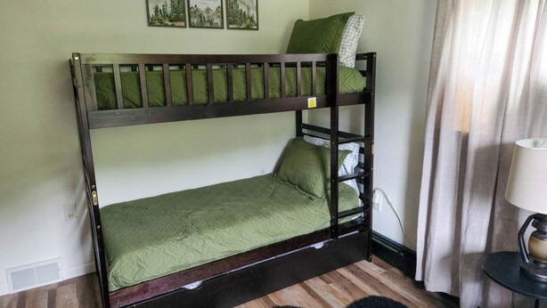 Una cama litera con ropa de cama verde, un cuadro colgado en la pared, una lámpara y una cortina.