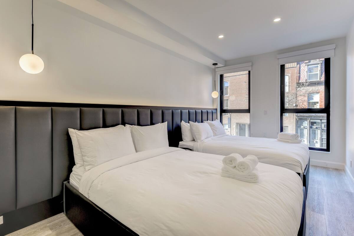 A modern hotel room with two beds, a dark headboard, and a view of the city through large windows.