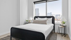 Een moderne slaapkamer met een zwart metalen bed en witte beddengoed, met een stadszicht door grote ramen.