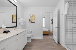 Un baño moderno con un espejo grande, un lavamanos y un inodoro. Se ve una cama al fondo.