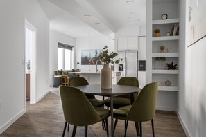 Una cocina moderna con armarios blancos, un comedor con una mesa redonda y sillas verdes, y un gran ventanal que proporciona luz natural.