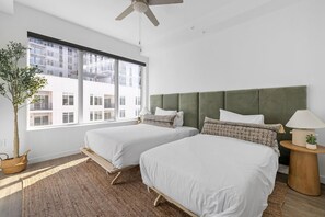 Un dormitorio moderno con dos camas, un ventanal grande, un ventilador de techo y vistas a un edificio.