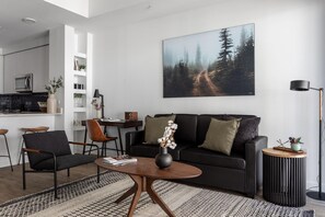 Una sala moderna con un sofá oscuro, dos sillones, una mesa de centro y un cuadro de un bosque en la pared.