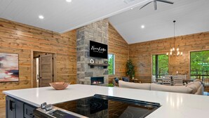 A modern kitchen with a stone fireplace, a flat-screen TV, and a large island with a built-in stovetop. - Hot Tub | Fire Pit | Corn Hole (Broken Bow)