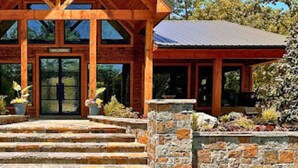 A wooden lodge with a prominent gable roof, a stone entrance, and a covered porch area. - Hot Tub | Fire Pit | Corn Hole (Broken Bow)
