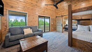 A modern log cabin interior with a sofa, a wooden coffee table, a bed, and a ceiling fan. - Hot Tub | Fire Pit | Corn Hole (Broken Bow)