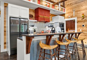 A kitchen with white cabinets, a red upper cabinet, a stainless steel refrigerator, a microwave, and wooden stools.