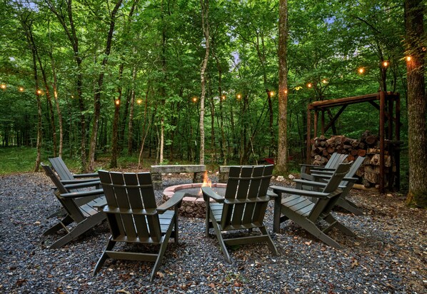 A forest setting with a fire pit and Adirondack chairs.