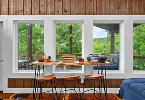 Una habitación con una mesa de madera, taburetes de bar y un sofá. Hay tres ventanas con vistas a zonas verdes.