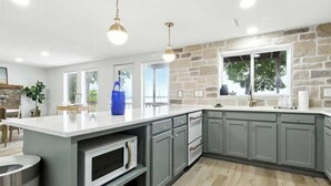 A modern kitchen with grey cabinets, a white island, and a stone accent wall. - Hot Tub, Main Channel Views, Great Outdoor Spaces, Blackstone (Sunrise Beach)