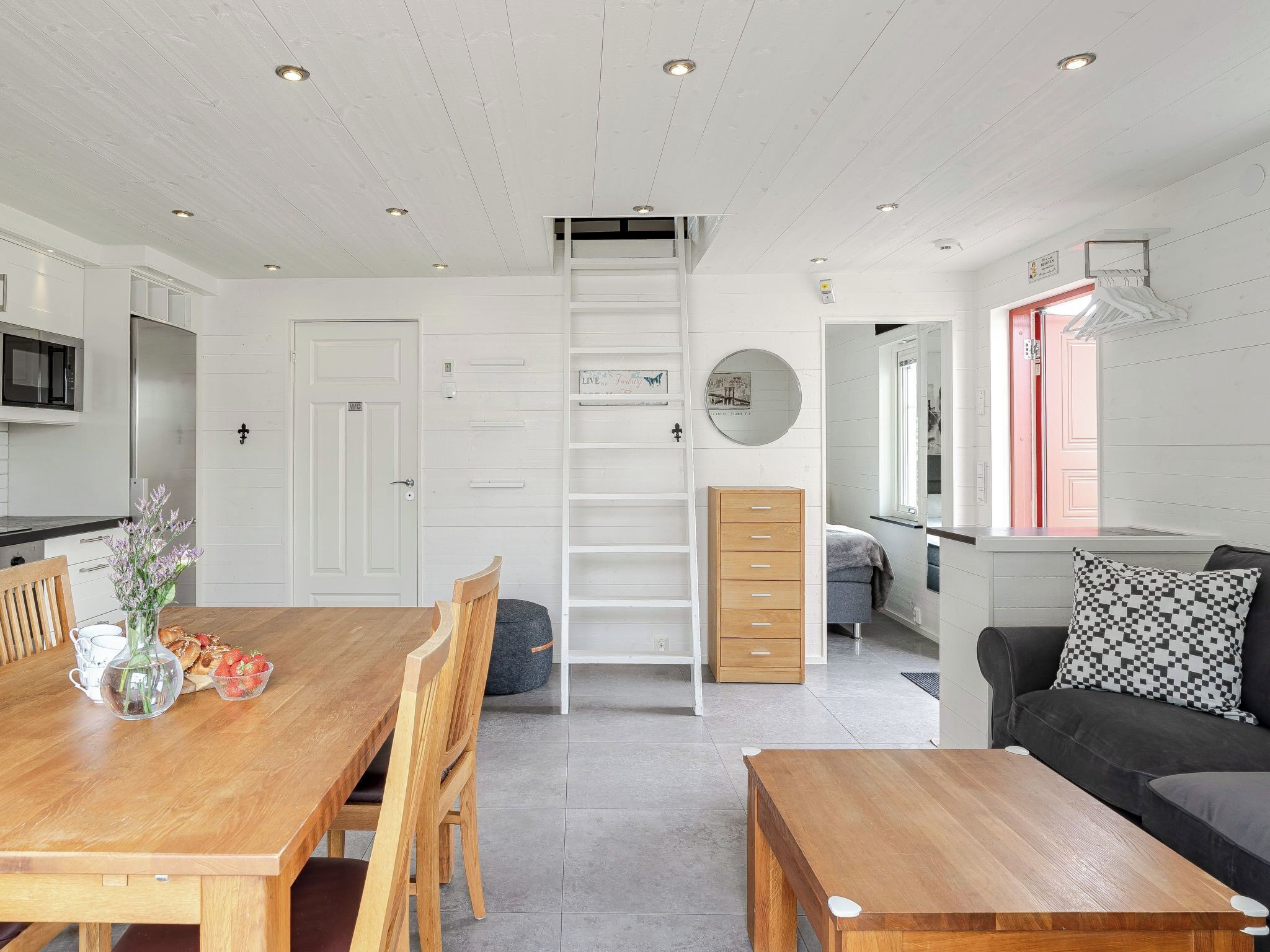A modern kitchen with white cabinetry, a wooden dining table with chairs, a built-in ladder, and a black sofa with a patterned pillow.