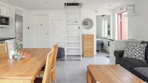 A modern kitchen with white cabinetry, a wooden dining table with chairs, a built-in ladder, and a black sofa with a patterned pillow. - Östra Stranden by Interhome (Halmstad)