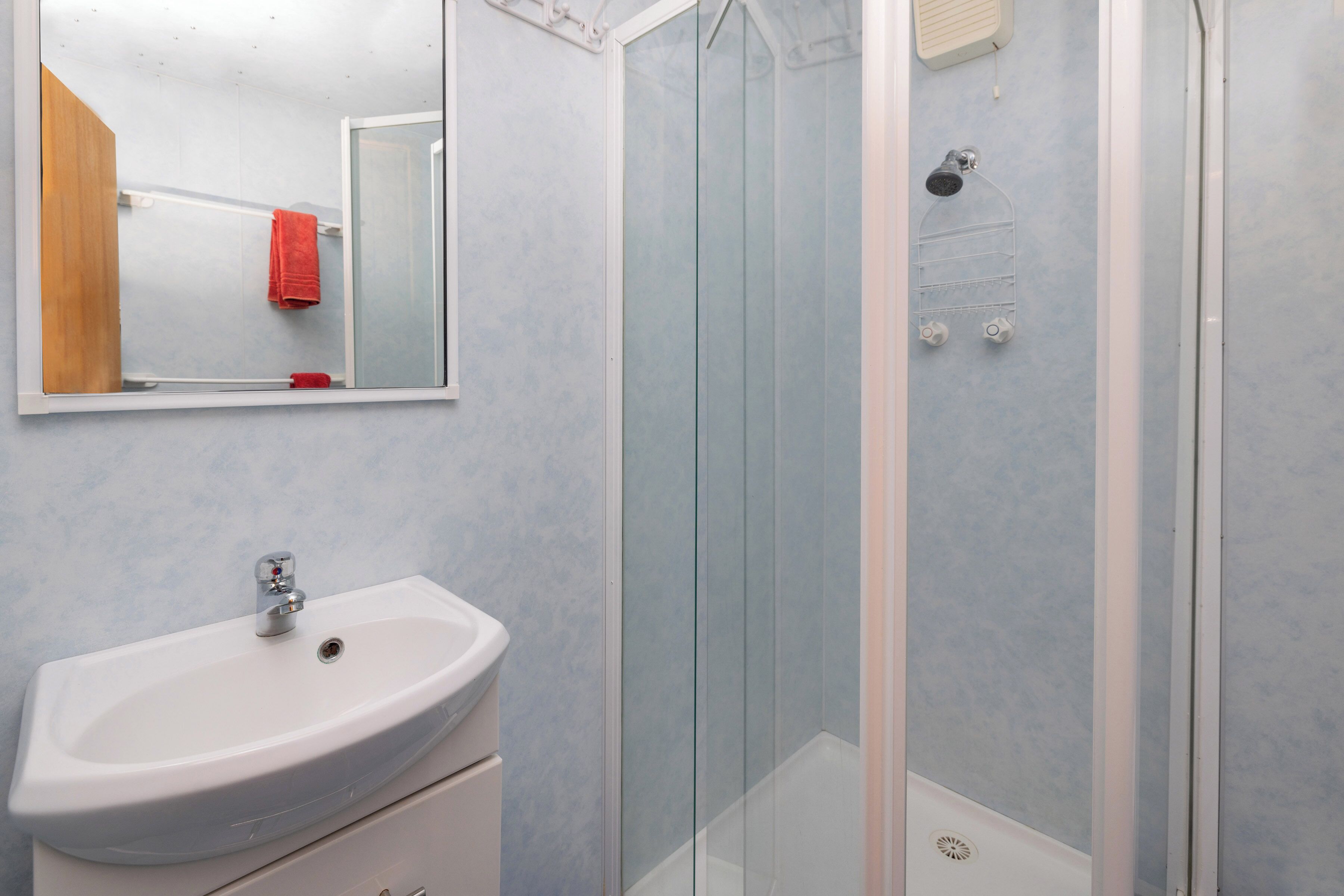 Une salle de bain avec un lavabo blanc, une cabine de douche en verre et un miroir qui reflète une serviette rouge.